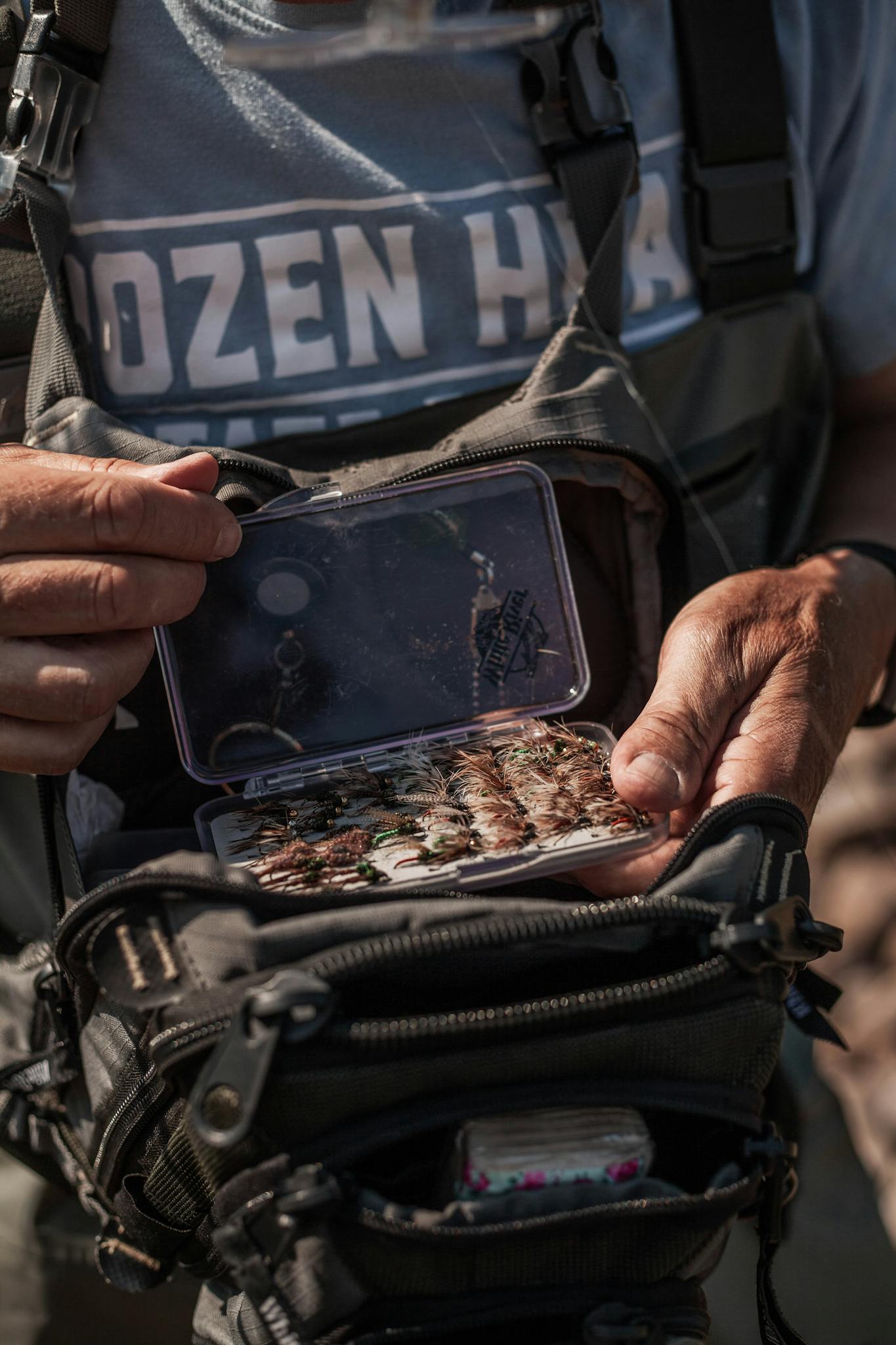 A man prepares his fishing tackle by a river, organizing equipment for outdoor fishing.